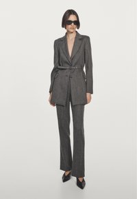 Frau in einem maßgeschneiderten dunkelgrauen Blazer mit Gürtel und passender Hose, schwarzen spitzen High Heels und übergroßen schwarzen Sonnenbrillen, die vor weißem Hintergrund steht.