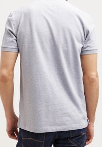 Muž v jednoduchém světle šedém polo tričku s krátkým rukávem a tmavě modrých džínách, zobrazený zezadu na bílém pozadí.