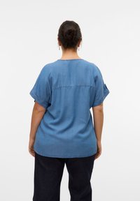 Woman with dark hair in a bun wearing a loose-fitting blue short-sleeve shirt and dark pants, standing facing away against a white background.