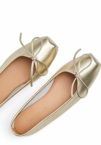 Bailarinas doradas con punta redondeada y acabado metálico. Presentan un pequeño detalle de lazo en la parte delantera y un interior suave.
