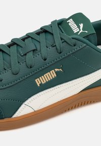 Sneaker verde in pelle con suola in gomma bianca e beige. Presenta il logo "PUMA" in oro, una superficie testurizzata e lacci multicolori.