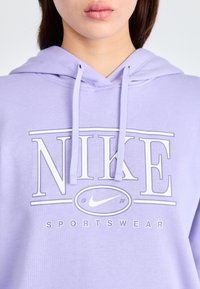 Haut à capuche violet clair avec un graphique sur le devant affichant "NIKE" et "SPORTSWEAR" en blanc. Comprend des cordons de serrage et une poche kangourou.
