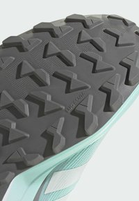 Podeszwa zewnętrzna buta trailowego wykonana z czarnej gumy o wyraźnej fakturze i agresywnym bieżniku, uzupełniona przez jasną miętową cholewkę i białe detale.