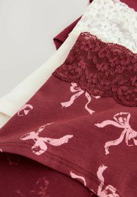 Gevouwen bordeauxkleurige stof met roze strikpatronen en bordeauxkleurige en witte kantafwerking op een lichte achtergrond.