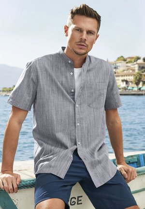 Graues kurzärmliges Hemd mit Leinenstruktur, Stehkragen und Brusttasche, kombiniert mit dunklen Shorts. Im Hintergrund sind Wasser und Gebäude zu sehen.