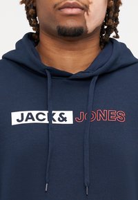 Felpa blu navy in cotone con tasca frontale. Presenta "JACK&" in bianco e "JONES" in rosso, con stringhe e una texture morbida.