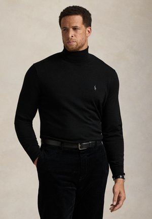 Man with short curly hair and beard wearing black turtleneck sweater, black pants, and a black belt, standing against a light background.
