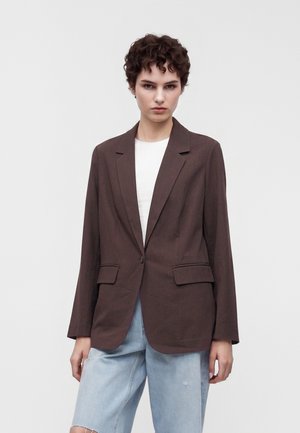 Jeune personne aux cheveux courts et bouclés porte un blazer marron foncé sur une chemise blanche et un jean bleu clair déchiré, debout devant un fond uni clair.