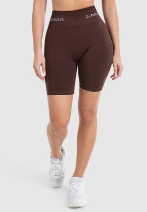 Shorts de vélo taille haute marron, tissu lisse, longueur mi-cuisse, avec une bande de logo blanche contrastante à la taille, associés à des sneakers blanches.