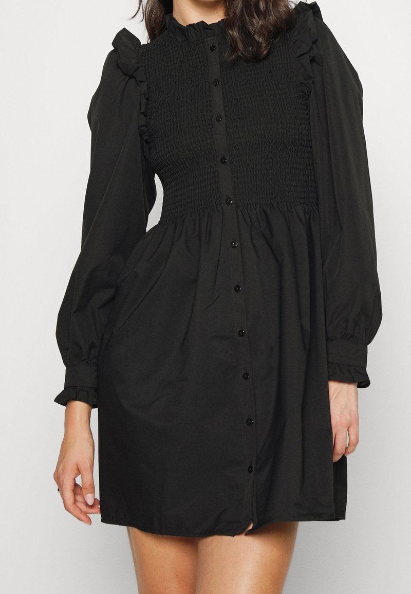 Femme portant une mini-robe noire à manches longues avec un corsage smocké, des épaules et poignets volantés, et une fermeture avec boutons à l'avant.