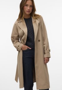 Trench coat beige in tessuto morbido, con un design doppiopetto, ampie alette e tasche laterali. Abbinato a un maglione blu navy e jeans scuri.