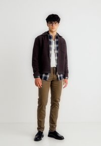 Νεαρός άνδρας με γυαλιά φορώντας στρωμένο καφέ σακάκι, πουκάμισο με καρό σχέδιο, λευκό t-shirt, χακί παντελόνι και μαύρα παπούτσια, στέκεται μπροστά σε έναν απλό λευκό τοίχο.