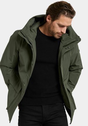 Man in een donkergroene hoodie over een zwarte gestructureerde sweater, kijkend naar beneden met zijn handen in de zakken van de jas tegen een effen achtergrond.