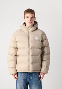 Young man wearing a beige puffer jacket with a hood and white CK logo, paired with blue denim jeans, standing against a plain background.