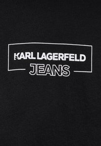 Juodas audinys su baltu logotipu, kuriame užrašyta "KARL LAGERFELD" viršuje ir "JEANS" apačioje, įrėmintas stačiakampio rėme. Tekstūra lygi.
