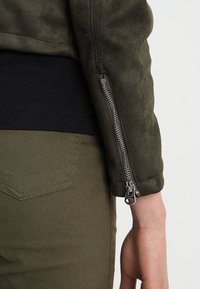 Giacca in suede verde oliva con dettagli della manica con zip, abbinata a una camicia nera e pantaloni verde oliva, con una tasca sul retro.