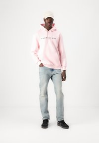 Hoodie roz cu logo-ul "TOMMY JEANS", blugi bleu deschis și pantofi negri. Modelul poartă o șapcă albă, stând în fața unui fundal neutru.