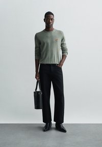 Lichtgroene trui met lange mouwen en geborduurd logo, zwarte wijde broek, zwarte schoenen en een cilindrische tas met schouderriem.