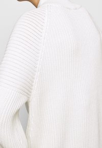 Pull en tricot blanc avec une texture côtelée, un col montant et un design à épaule déchue. Le tissu semble doux et léger.