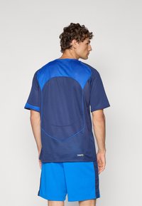 Tricou sportiv de marină cu accente de plasă albastră pe umeri și pe spate. Mâneci scurte, gât rotund, material ușor și margini curbe.