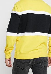 Žlutá mikina s černobílými vodorovnými pruhy, vyrobená z měkké látky s žebrovanými manžetami a pasem, pohodlný střih.