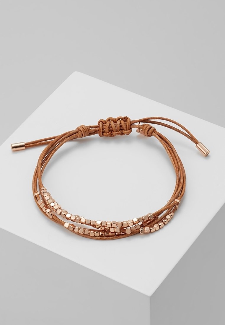 Bracelet en cuir brun multi-brins avec de petites perles dorées en forme de cube et une fermeture nouée réglable, présenté sur un socle blanc en forme de cube.