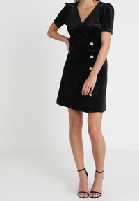 Czarna sukienka kopertowa z miękkiej tkaniny, z krótkimi bufkami na rękawach, głębokim dekoltem w serek oraz ozdobnymi guzikami z przodu.