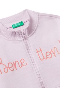 Giacca lilac con zip, colletto a costine e texture morbida, con la scritta in glitter rosa "Boene" sulla parte anteriore. Etichetta verde del marchio visibile.