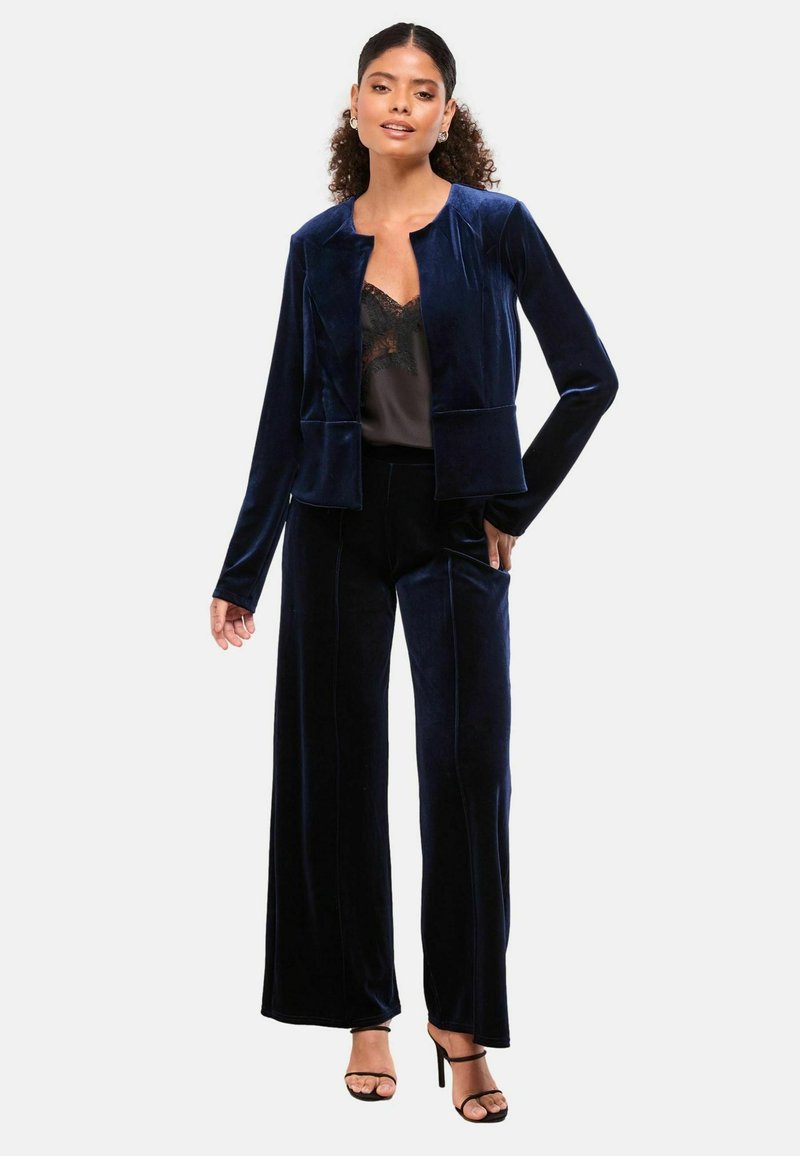 Navy tamni baršunasti odijelo koje se sastoji od prilagođene jakne s okruglim izrezom i širokim hlačama. Cjelina ima glatku teksturu i suptilan sjaj.