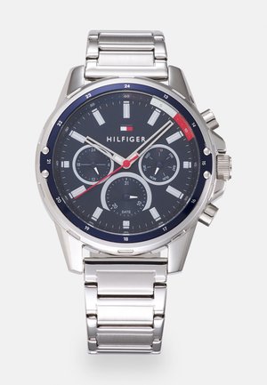 Silver stainless steel watch with a dark blue dial, featuring three sub-dials, white hour markers, and red accents. Model name "HILFIGER" displayed.