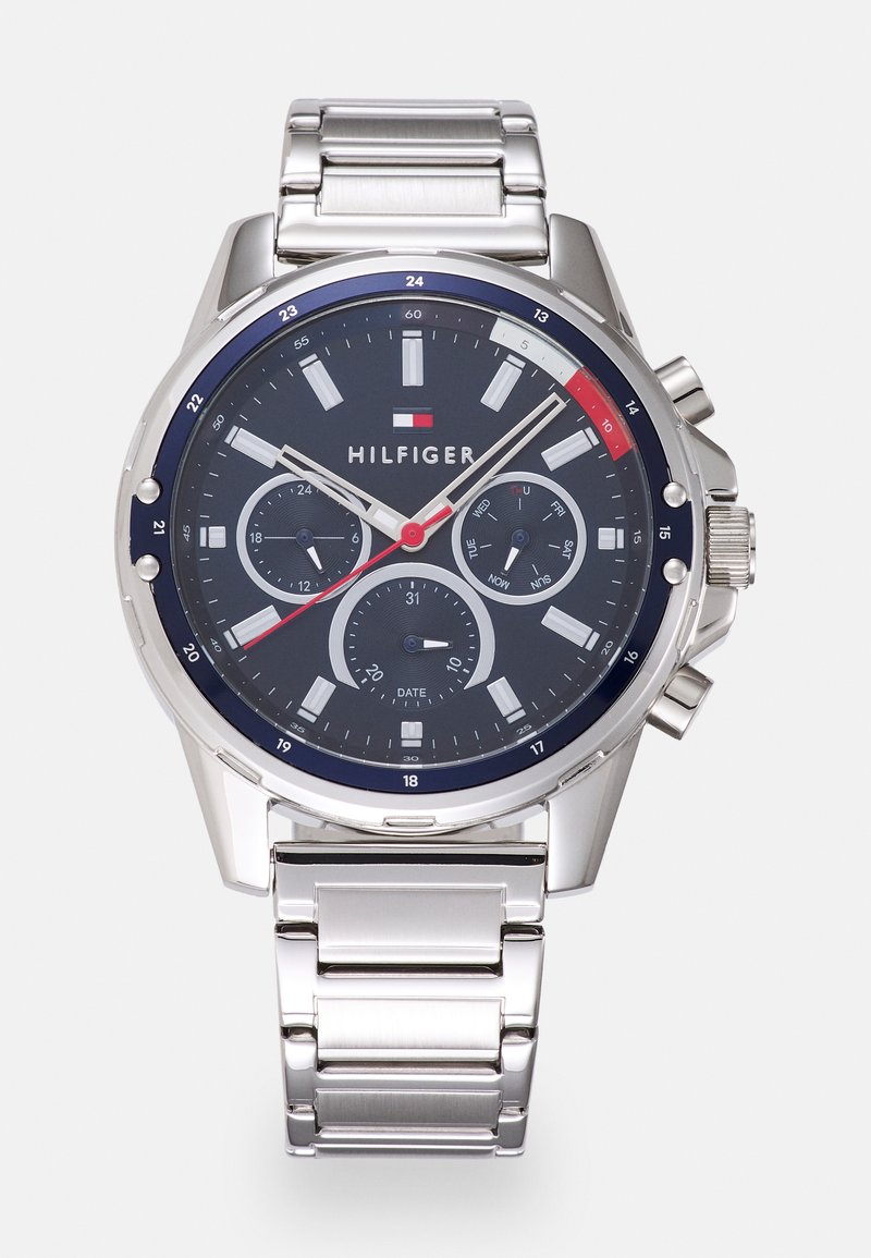 Sølv rustfritt stålklokke med en mørk blå skive, med tre subskiver, hvite timemarkører og røde detaljer. Modellnavn "HILFIGER" er vist.