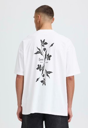 Homme portant un T-shirt blanc surdimensionné avec un motif floral noir et le texte "Solid EST. 1999" au dos, associé à un jean foncé.