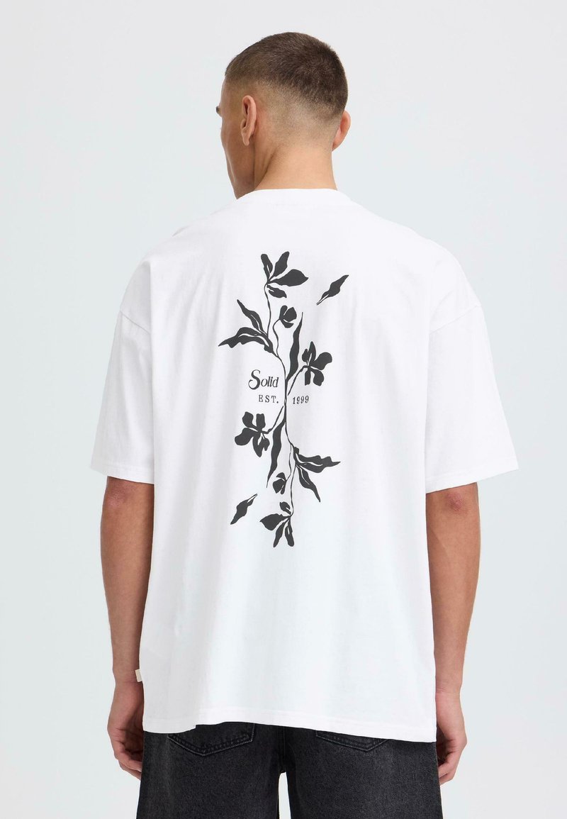Homme portant un T-shirt blanc surdimensionné avec un motif floral noir et le texte "Solid EST. 1999" au dos, associé à un jean foncé.