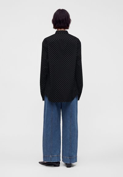 Personne aux cheveux courts et foncés se tient de dos, portant une chemise noire à manches longues à motifs et un jean bleu à jambes larges avec des chaussures noires.
