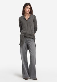 Graue Knopfleiste Strickjacke aus weichem Stoff, kombiniert mit hellgrauen Jeans mit weitem Bein. Lockerer Schnitt mit gebundenem Detail in der Taille. Flache Schuhe.