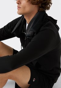 Felpa nera con zip indossata con un asciugamano nero drappeggiato sulle spalle, abbinata a pantaloni corti sportivi neri con logo. Stoffa dalla texture liscia.