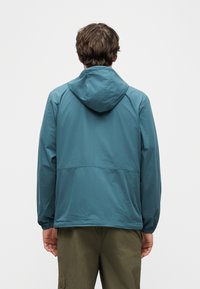 Άνδρας που φοράει γαλάζιο μπουφάν με κουκούλα και χακί παντελόνι, εμφανιζόμενος από πίσω, στέκεται μπροστά σε απλό λευκό φόντο.