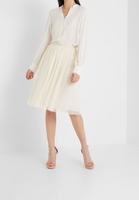 Blouse à manches longues crème avec détails à boutons, associée à une jupe en tulle crème longueur genou avec un motif à pois, portée avec des talons nus.