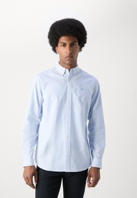 Lysblå button-up skjorte med smalle hvide striber. Har en krave, lange ærmer og et lille logo på venstre bryst. Glat tekstur.