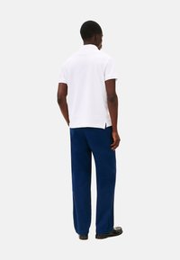 Polo shirt bianco in cotone a maniche corte, abbinato a pantaloni blu a gamba larga. Presenta spacchi laterali all'orlo. Scarpe nere visibili.
