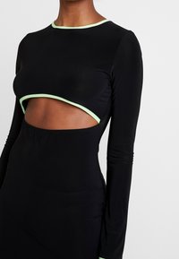Robe noire à manches longues, avec une découpe au niveau du ventre et des accents vert clair le long de l'encolure et du bord découpé. Texture de tissu lisse.