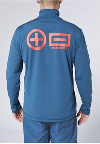 Mann trägt blaue Langarmoberteil und Hose, Rücken zur Kamera gewandt, mit großen orangefarbenen Pluszeichen und Rechteckgrafiken auf dem oberen Rücken.