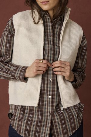 Femme portant une chemise à carreaux marron boutonnée et un gilet tricoté crème zippé, fermant la fermeture éclair du gilet avec les mains visibles, sur un fond marron.