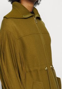 Olivgrön fleecejacka med ribbad krage, dragkedja och sidofickor. Har fransdetaljer längs ärmarna för extra textur.