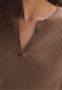 Zbliżenie na osobę w brązowej ryflowanej bluzce z dekoltem w literę V, odsłaniającą szyję i górną część klatki piersiowej.