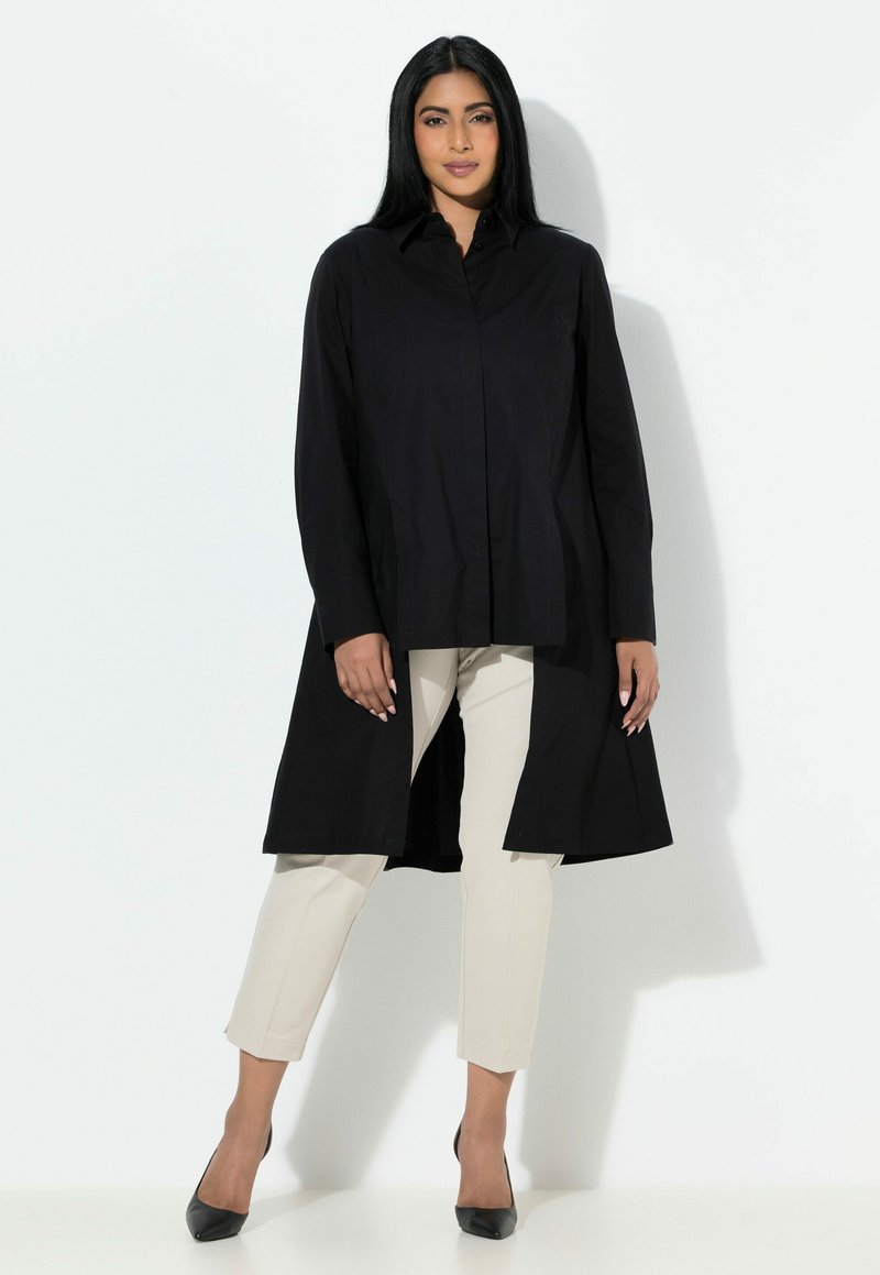 Czarna długa koszula z zapinanym na guziki przodem, zaokrąglonym dołem i długimi rękawami, w połączeniu z kremowymi spodniami i czarnymi szpilkami. Gładka faktura.