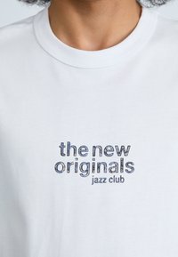 Vitt bomullströja med blå och svart grafisk text som säger "the new originals jazz club," med rund hals och korta ärmar.
