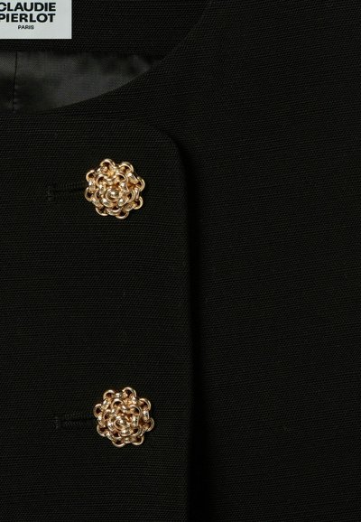 Veste en tissu noir avec deux boutons décoratifs en forme de fleur en métal doré, présentant un détail complexe et une doublure intérieure lisse.