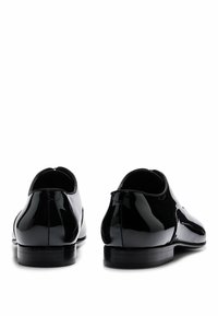 Chaussures en cuir verni noir avec une finition lisse, bout rond et petits talons noirs, présentant des coutures minimales et un design épuré.
