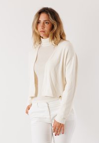 Donna con capelli biondi ondulati che indossa un dolcevita bianco sporco, cardigan e pantaloni, in piedi con una mano in tasca contro uno sfondo neutro.